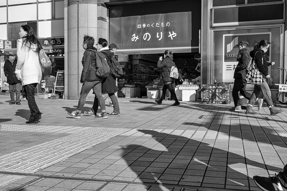 A monochrome low-angle photo of pedestrians passing by the entrance to the Tokyu grocery store next to Kanamachi Station in Tokyo. On the right of the frame in the foreground we see long shadows cast by people who are out of frame except for a portion of one foot clad in a New Balance sneaker. The grocery store entrance is in the rear center of the frame, with a line of four passing pedestrians demarcating the upper diagonal third of the frame. The produce section of the store is visible past the store entrance.