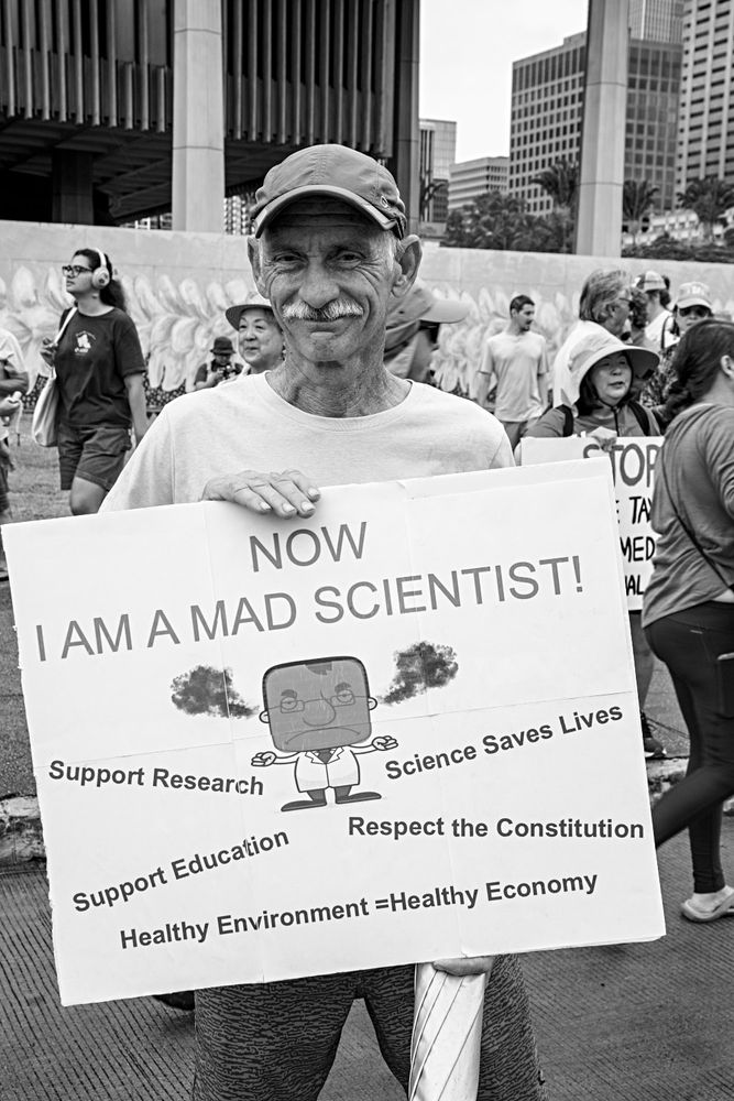 Monochrome portrait of an older man carrying a sign reading "NOW I AM A MAD SCIENTIST above a stylized imagine of a figure in a lab coat with smoke coming out of its ears. Below the figure are phrases reading "Support Research, "Science Saves Lives, "Support Education," "Respect the Constitution," and, finally, "Healthy Environment = Healthy Economy." In the background are other protestors and a posrtion of the decorated construction wall surrounding the Hawai'i state capitol building.