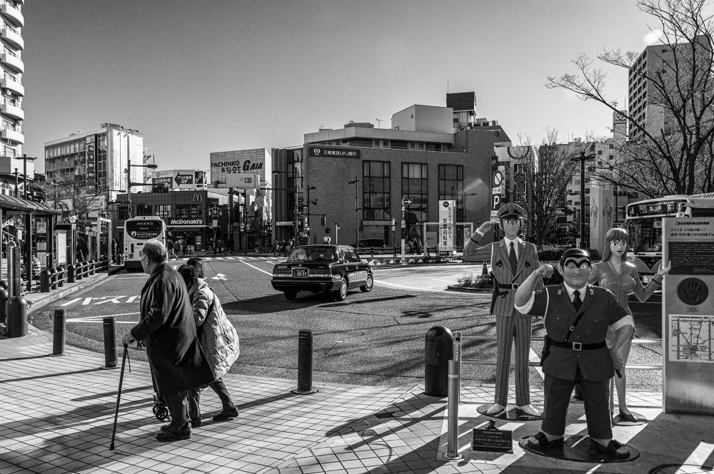 Monochrome photo outside Kameari Station in Katsushika, Tokyo. In the left foreground are an elderly couple making their way to the station entrance as the taxi in which they arrived pulls away. In the right foreground is a tableau featuring life-sized characters from a popular manga and anime series based in Katsushika, standing next to a map of the train lines. In the background are various buildings including an apartment block, a McDonald's, a pachinko hall and others. 