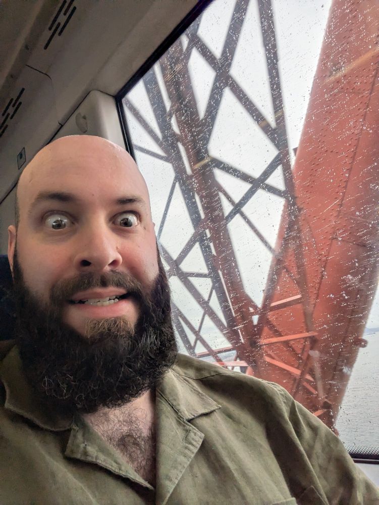 Matt crossing the Forth Bridge by train. The view out the window includes the waters of the Firth of Forth and the structure of the bridge itself, which is lots of steel struts painted a ruddy brown.