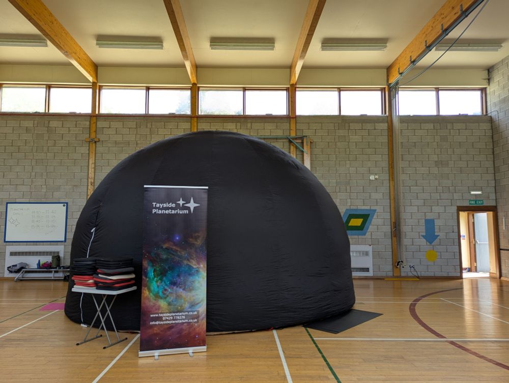 The portable planetarium set up in the gym hall at Morrison's Academy, Crieff. It is a big black hemispherical dome, 3.5m tall, with a table of cushions and the Tayside Planetarium banner in front of it.