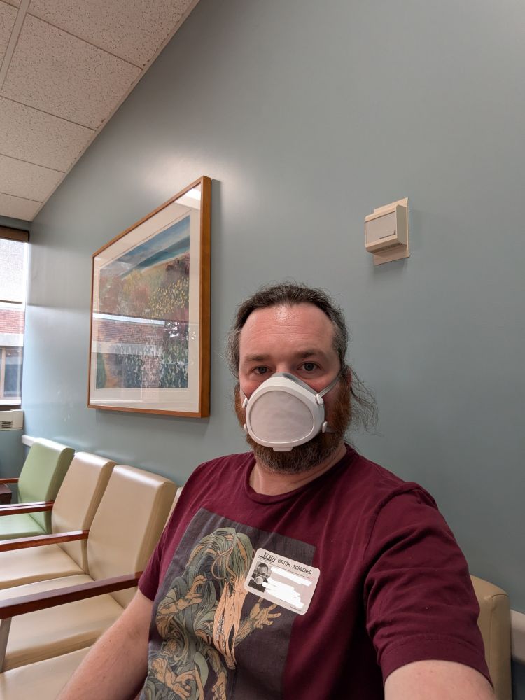 A white man with brown hair and beard, wearing a Flo mask, sits in an otherwise empty hospital waiting room.