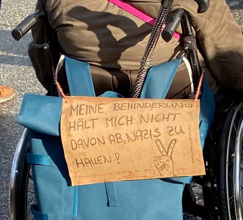 Eine ältere Dame im Rollstuhl mit einem Schild an der Rückseite auf dem steht: MEINE BEHINDERUNG HÄLT MICH NICHT DAVON AB, NAZIS ZU
HAUEN !