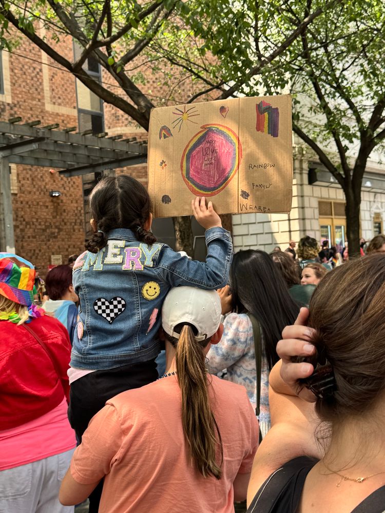 A little girl holding a sign that reads “More love, less hate” and “we are a rainbow family”