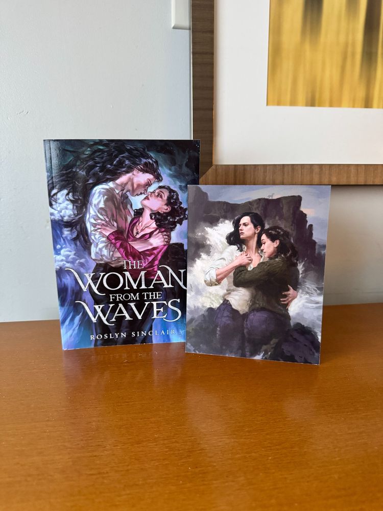 A photo of a paperback copy of Roslyn Sinclair's sapphic romance novel THE WOMAN FROM THE WAVES, standing on a table, next to a postcard with art of the two main characters.