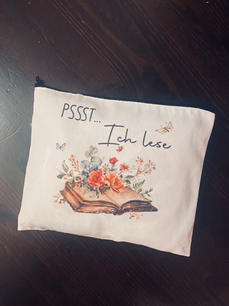 Beiges Stofftäschchen mit schwarzem Reißverschluss, darauf die Aufschrift ‚PSSST… Ich lese‘ in eleganter Schrift. Darunter eine Illustration eines aufgeschlagenen Buches, aus dem bunte Blumen und Zweige herauswachsen. Um die Illustration herum fliegen kleine Schmetterlinge.