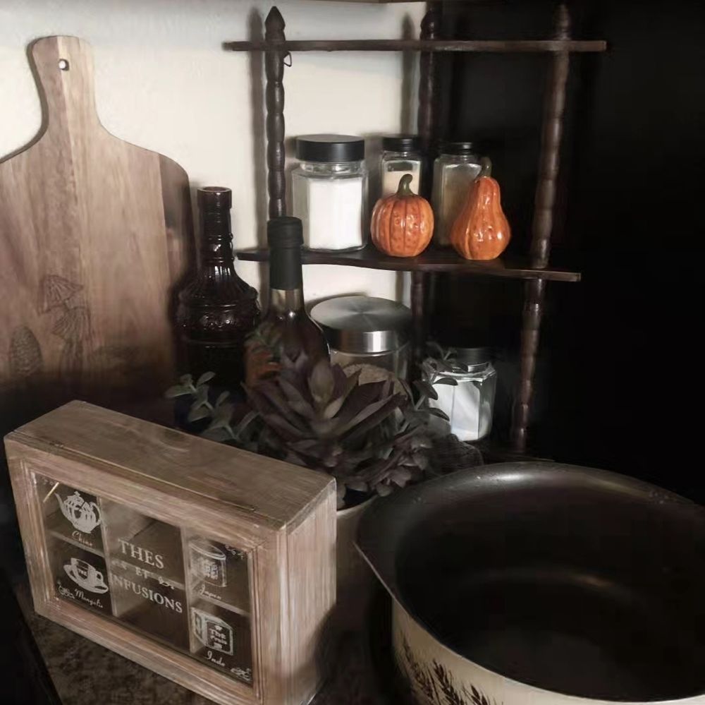 A photo of a kitchen counter with a small set of stand-up shelves in the corner. The shelves have multiple spices with a set of salt and pepper shakers shaped like pumpkins. A cutting board rests on the counter next to two bottles, an empty one and a non-alcoholic cider. In the foreground are a wooden tea box and some pots.