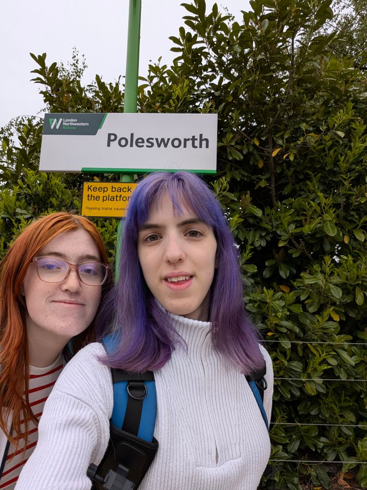 a picture of me and chloe stood underneath a polesworth station sign. the sign is white with green text. i have purple hair and i'm wearing a white jumper. chloe has ginger hair and is wearing a white and red stripy top. behind us (and the sign) is foliage. 
