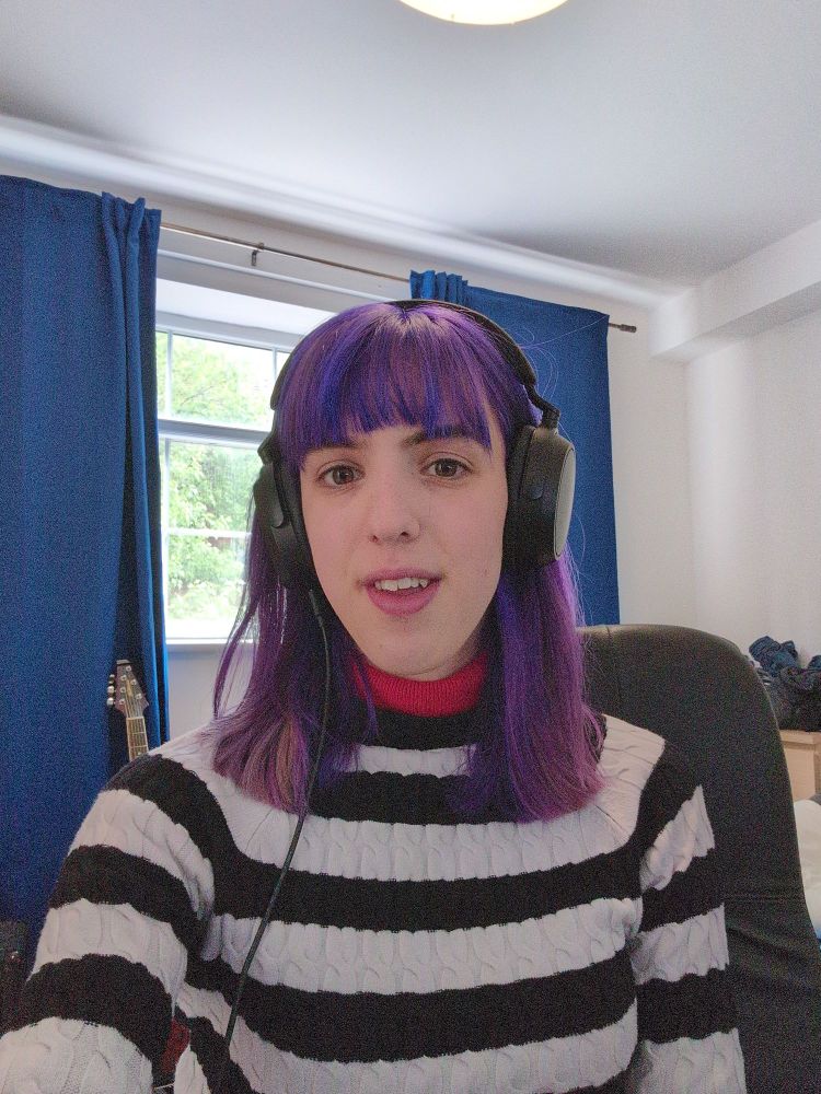 a selfie. i'm sitting in my bedroom. my hair is straight, purple, and shoulder length, with a fringe. i'm wearing a white and black striped jumper, with black over-ear headphones. behind me is the black desk chair i'm sat on, as well as the top of my guitar and my half-open blue curtains.