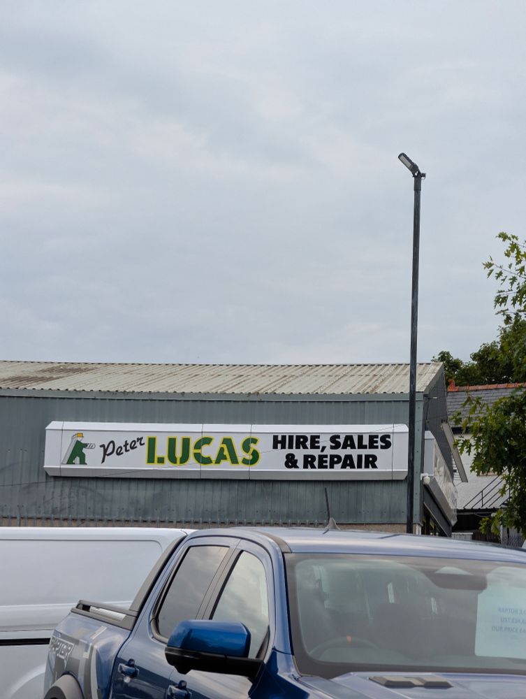 a sign on a warehouse reading "Peter LUCAS HIRE, SALES & REPAIR". the sign is white. the text has 3 different styles. the sky above is grey and cloudy, and in front the warehouse the top half of a blue pickup truck is visible.