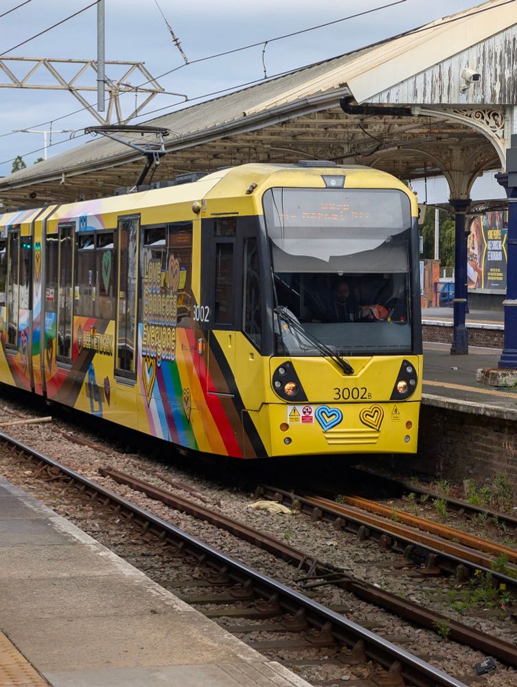 a picture of metrolink's pride vehicle, number 3002. it's in a bright yellow livery, with rainbows draped across the sides, partially covered by text reading "Love Connects Everyone". There's a blue and a yellow heart on the front. behind the vehicle is one of the platforms at altrincham station, with a historic cream canopy overhead.