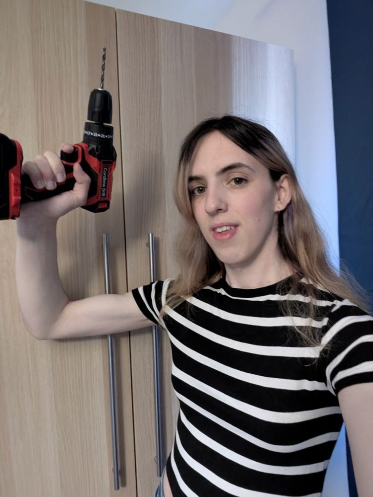 a selfie. i am holding a red and black drill. i'm wearing a black and white stripy short sleeve top. i have long hair that is currently an interesting mix of blonde, brown, and purple. i'm standing in front of my wardrobe, which has 2 newly installed tall metal handles attached to the doors. 