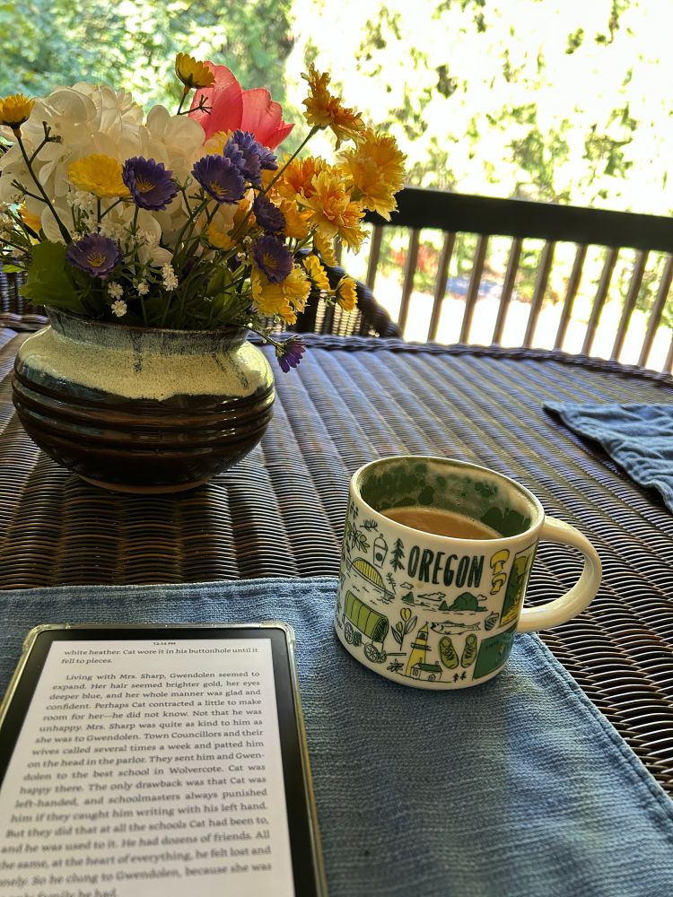 A Kindle and a coffee cup sitting on a blue placemat on an outdoor table. Behind them is a bowl of fake wildflowers. You can see sun and green trees in the background.