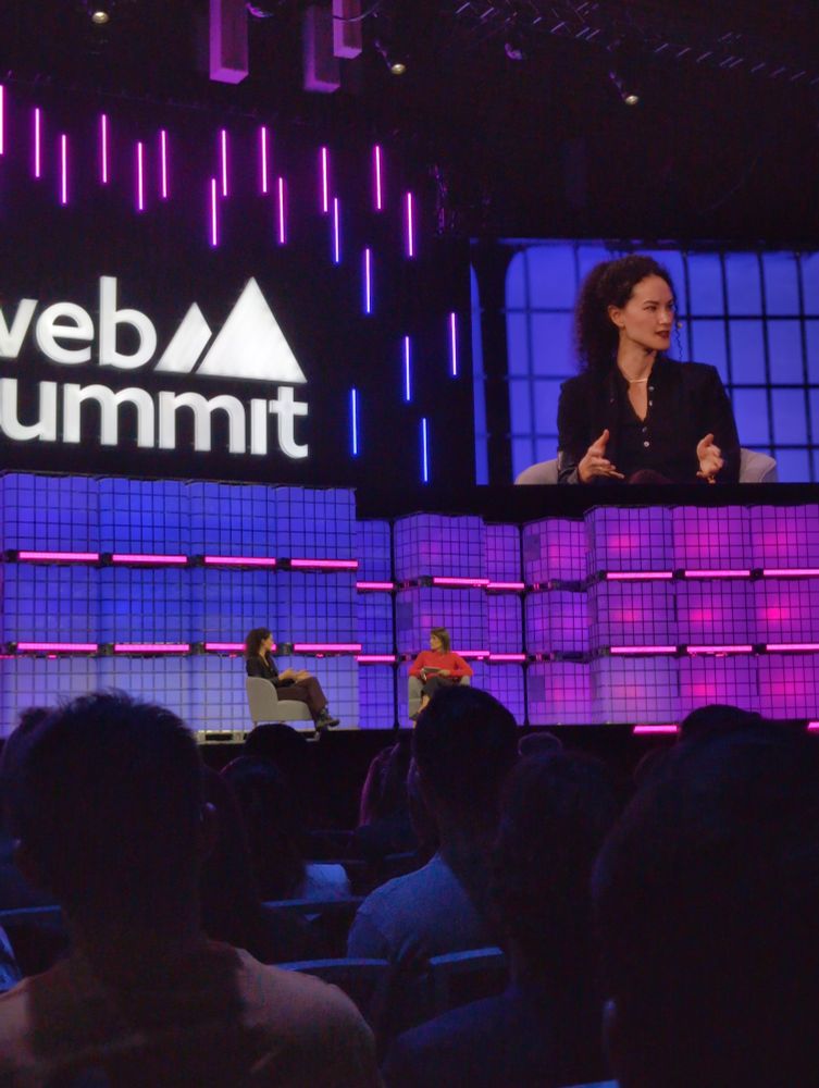Jay Graber and Katie Drummond talking on the main stage at Web Summit Vancouver, photo taken over the heads of other attendees, with a background to the subjects in neon pinks and blues