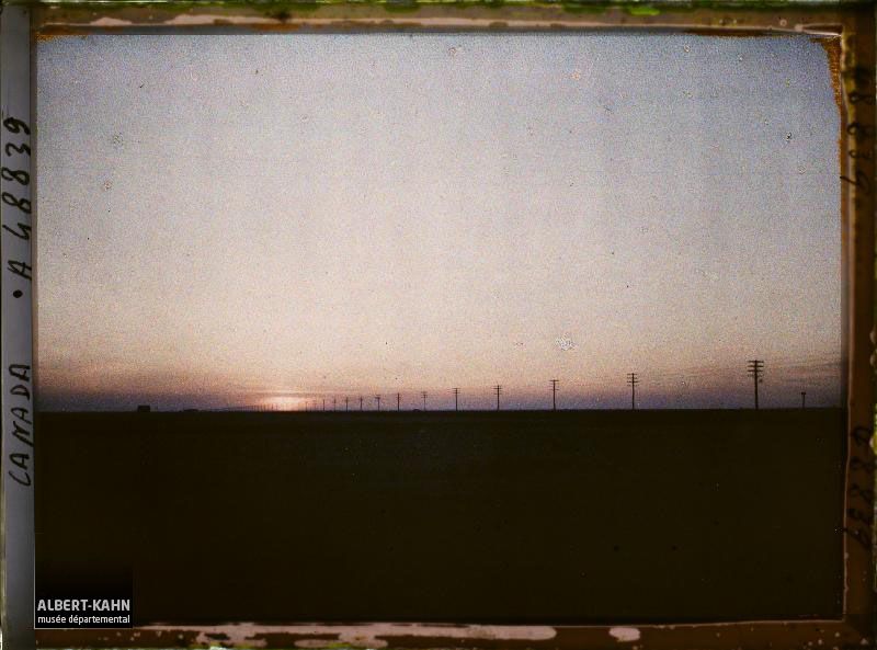 Sunset, the sky yellow blue fringed by clouds on the far images. Everything below the horizon is black, the silhouette of a building discernible to the left. From the center left and verging to the foreground on the right, a line of power poles strung with eight sets of wire, hardly distinguishable against the sky. Photo by Frédéric Gadmer, taken in Canada, 28 April, 1926.  