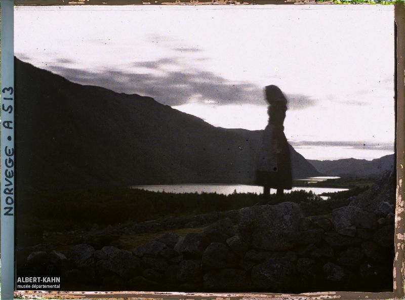A young woman stands on a stone. She faces left and wears a long black dress with a white apron. Her head is in motion. Little else can be discerned as she stands before a fading sun over a barren valley, all enveloped in shadow, a sky streaked with cloud reflected in water fringed by dark trees tapering off into the far reaches of the valley. Photo by Auguste Léon, captured 8 September, 1910, Près de Dalen, Norway.