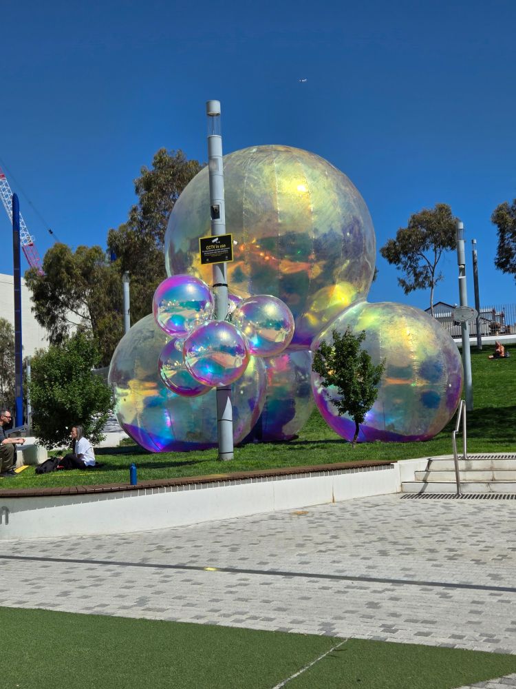 Smaller pearlised bubbles affixed to a light pole with bigger bubbles in the background, all on a grassy slope. 