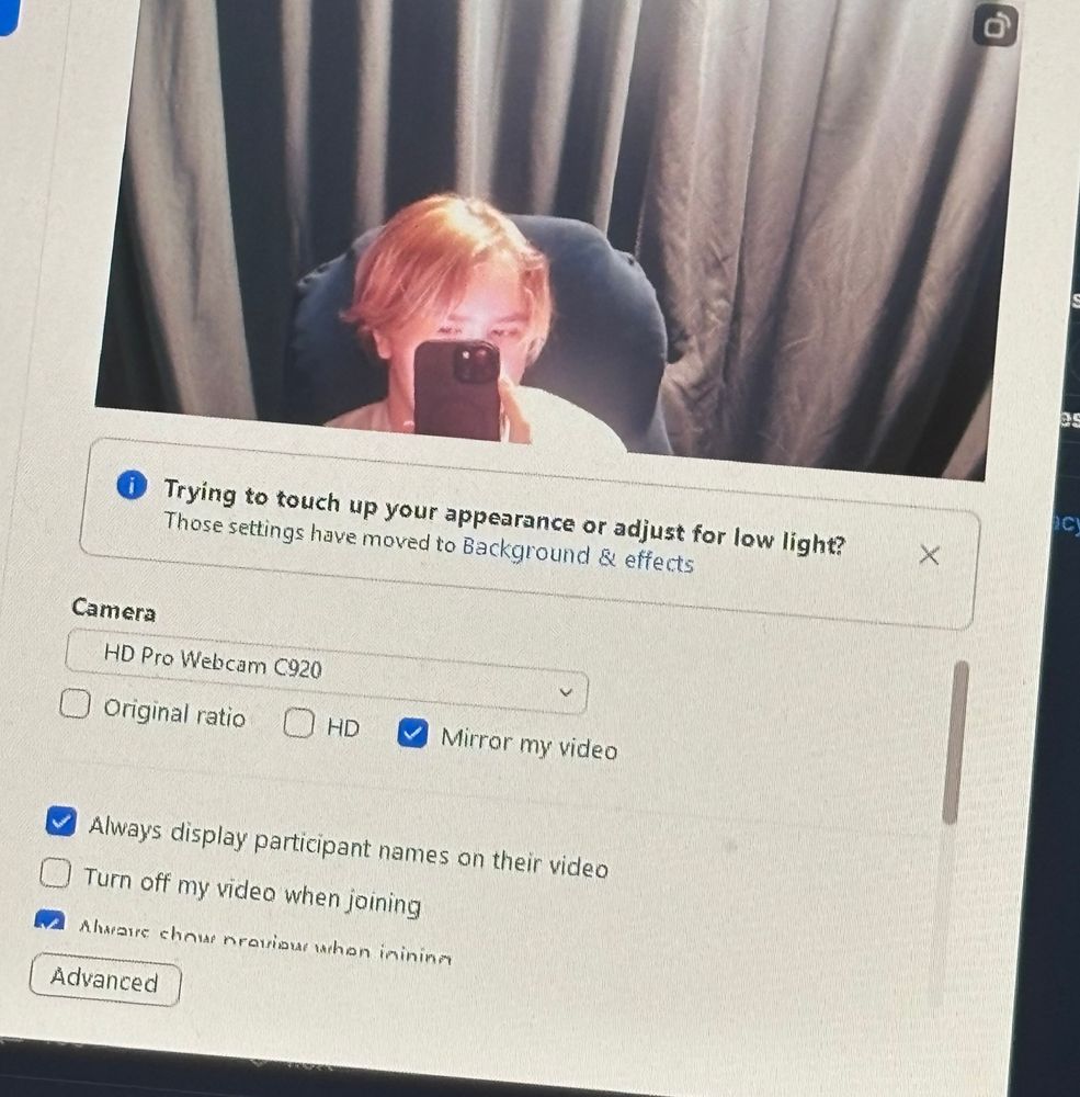 photo of zoom camera settings screen with person sitting in a black chair with black curtains seen behind them; the person has short hair and is holding a black phone