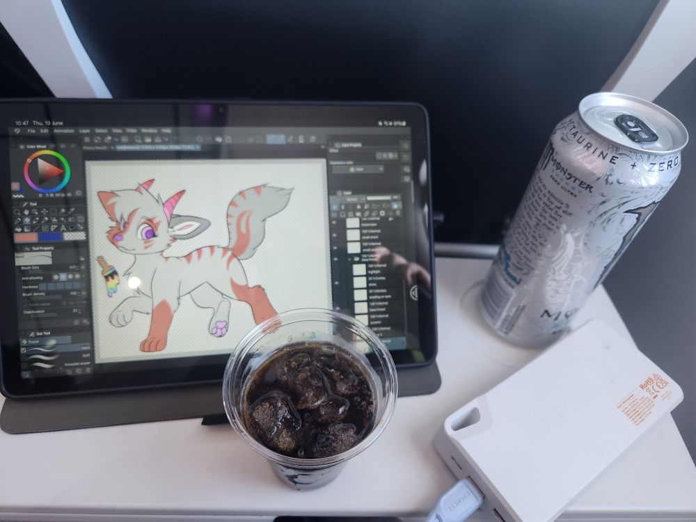 A picture taken inside a plane,shown is a alcoholic cola beverage,white monster,a power bank and a tablet with a unfinished project on screen,all resting on a tray table