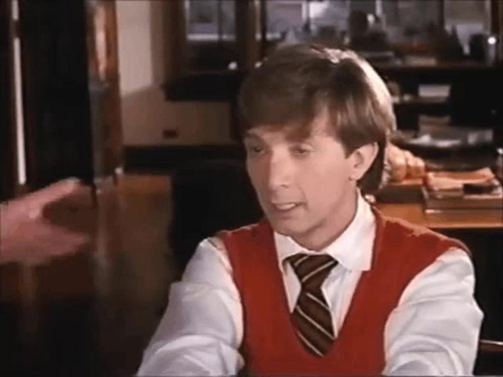 a man in a red vest and tie is sitting at a table talking to another man .