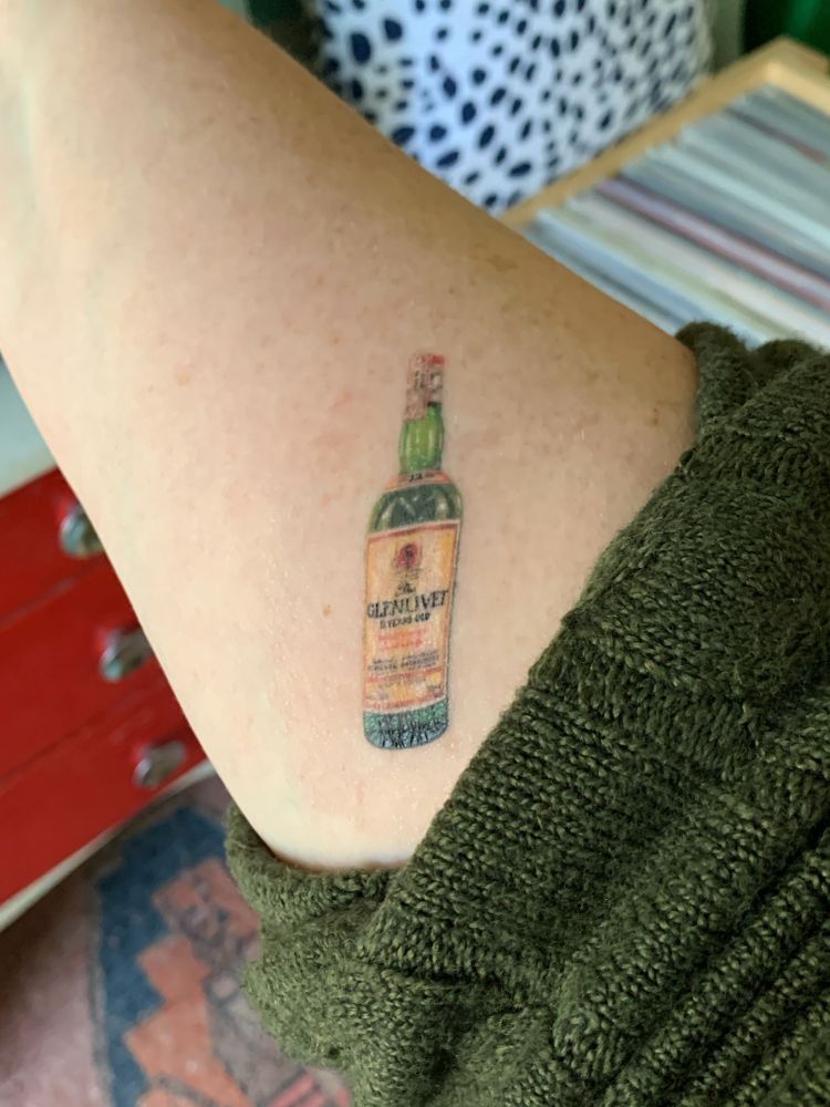 Pic of an extended right forearm, green sweater sleeve pushed up, revealing a small, delicately done, colour tattoo of a 1980s Glenlivet scotch bottle. 