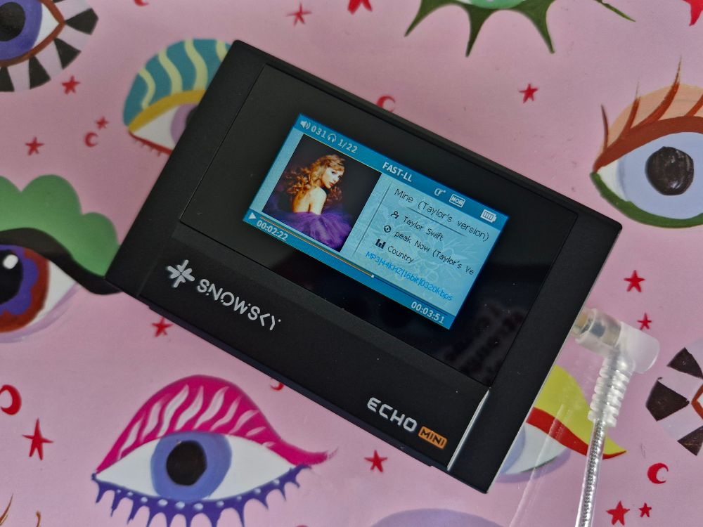 Photograph of a rectangular MP3 player, the Snowsky Echo Mini, with headphones plugged in. The display is currently on, displaying the currently played song, Mine (Taylor's Version) from the album Speak Now (Taylor's Version) by Taylor Swift.