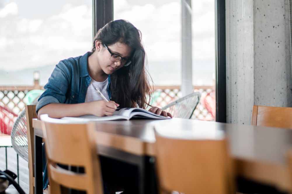 Student with long brown hair and glasses looks over and writes in book and a long table.