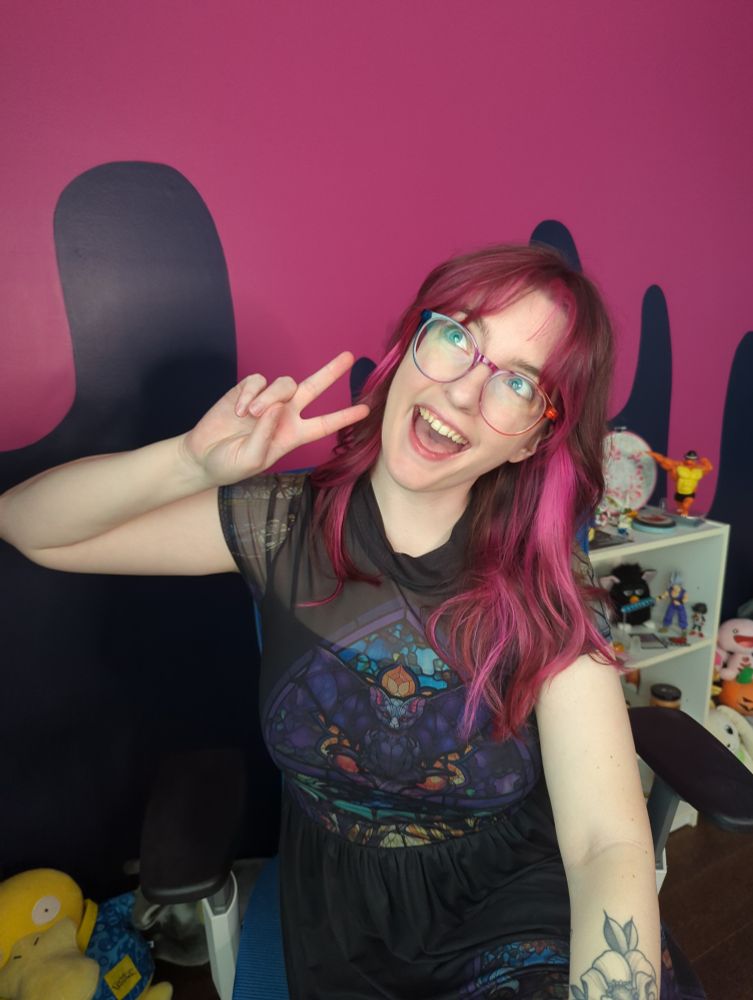 A selfie of Harpy making a silly face. She is wearing a dress with a bat stained glass design. She has pale skin, glasses, and medium length brown hair with curtain bangs and pink pieces in the front. Her stream room wall is navy blue with a pink drip design 