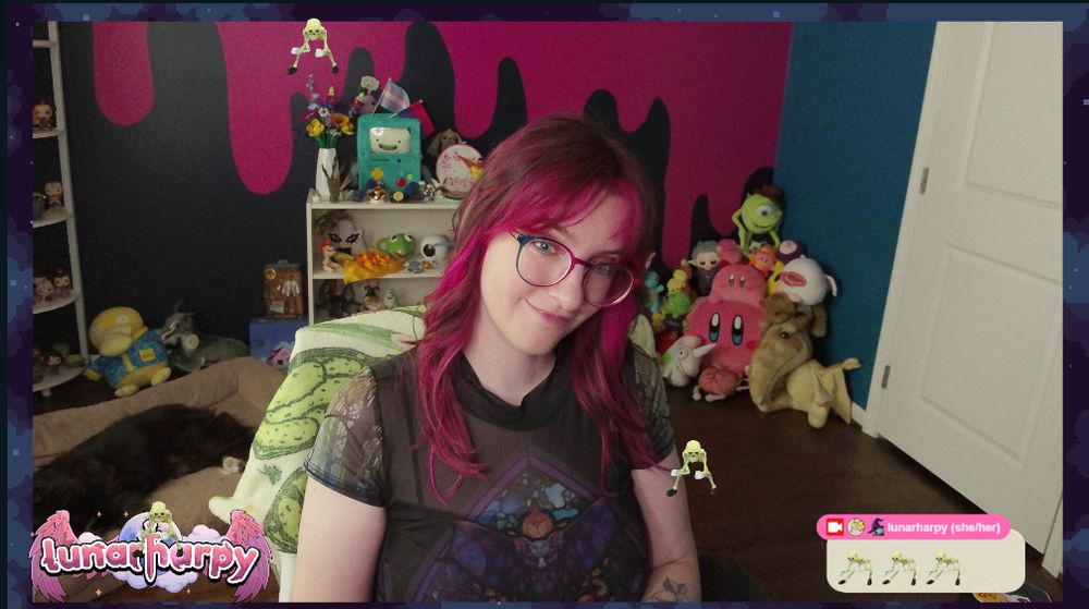 A webcam snapshot of Harpy in her stream room wearing a dress with a mesh top and a bat stained glass design. She has pale skin, glasses, and medium length brown hair with curtain bangs and pink pieces in the front.