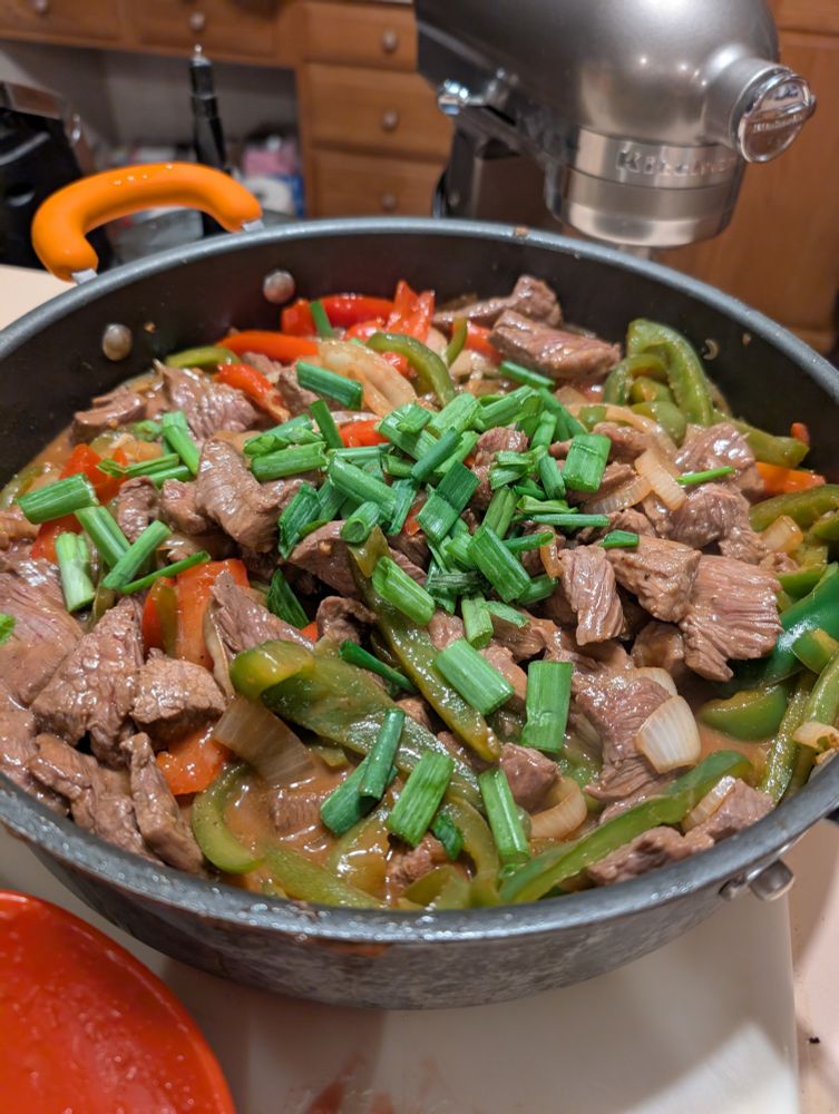 A large saute pan filled with steak pieces, red bell peppers, green bell peppers, onions and topped with scallions