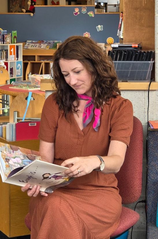 A woman in a brown dress and pink neckscarf reading Enough Is..., a picture book. 