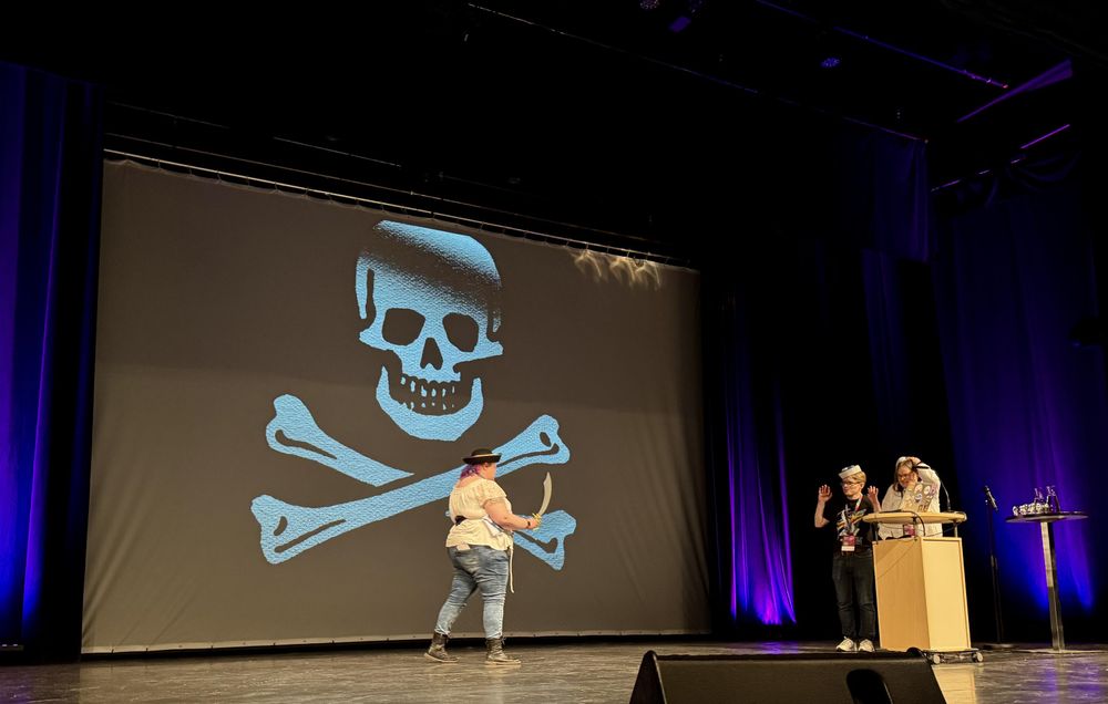 A pirate-themed presentation on stage features a large skull and crossbones backdrop. A person dressed in pirate attire with a sword walks toward two others standing beside a table, raising their hands in a playful manner.