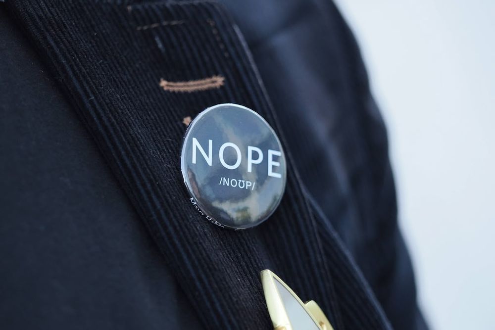 A closeup of a button, worn on a black jacket. The button is black and has the word “NOPE” in white text, along with a phonetic pronunciation guide.