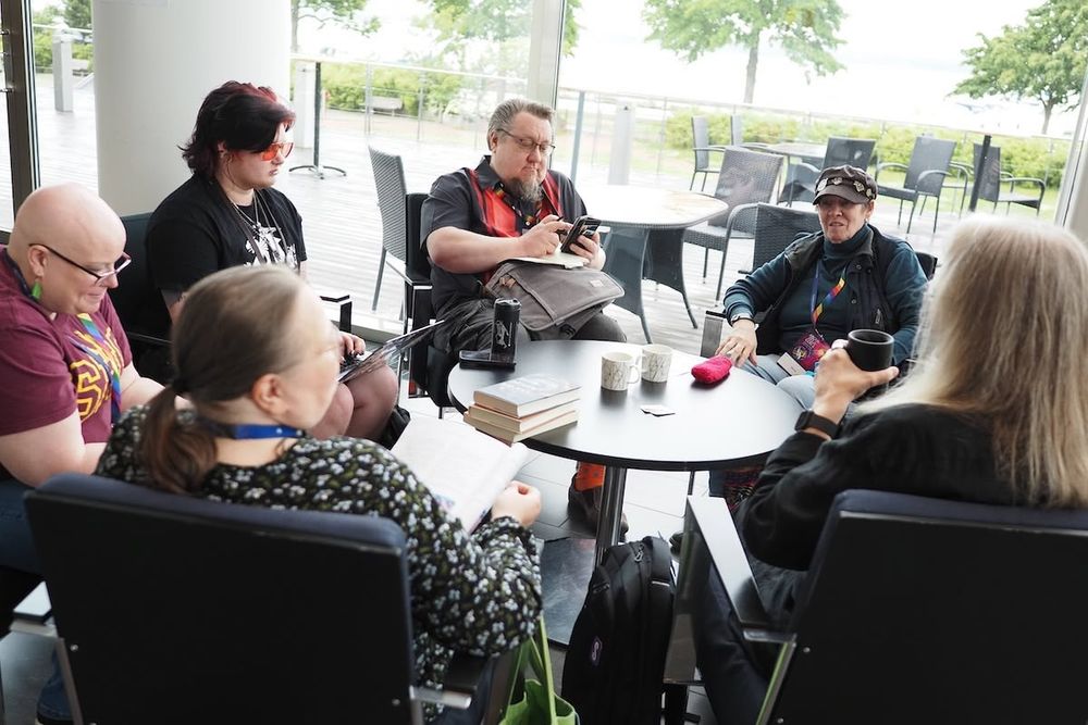A group of people is seated around a small table in a cafe setting. They are engaged in conversation, one person using a laptop and another their phone. The background features greenery and outdoor seating. On the table, there are books and coffee mugs.