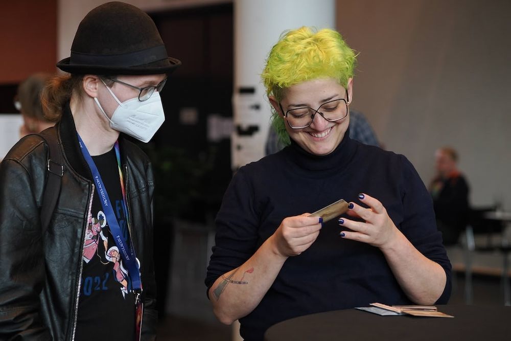 A person with large glasses is looking at a card and laughing. Another person in a hat and a mask is standing beside them, also looking at the card.