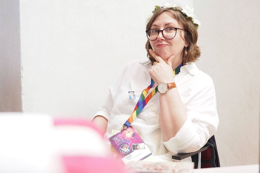 A woman dressed in a white shirt, wearing glasses with dark rims. Hand on chin, looking at the camera with a smile.