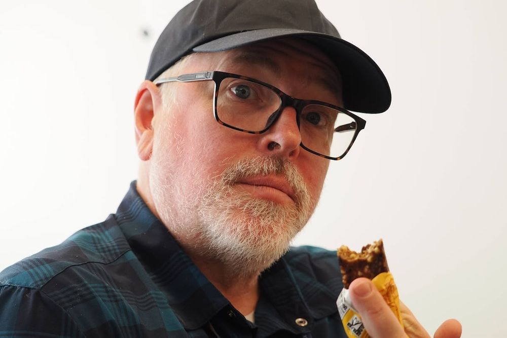 A closeup of a man with light grey beard, wearing black-rimmed glasses and a dark baseball cap, looking directly in to the camera