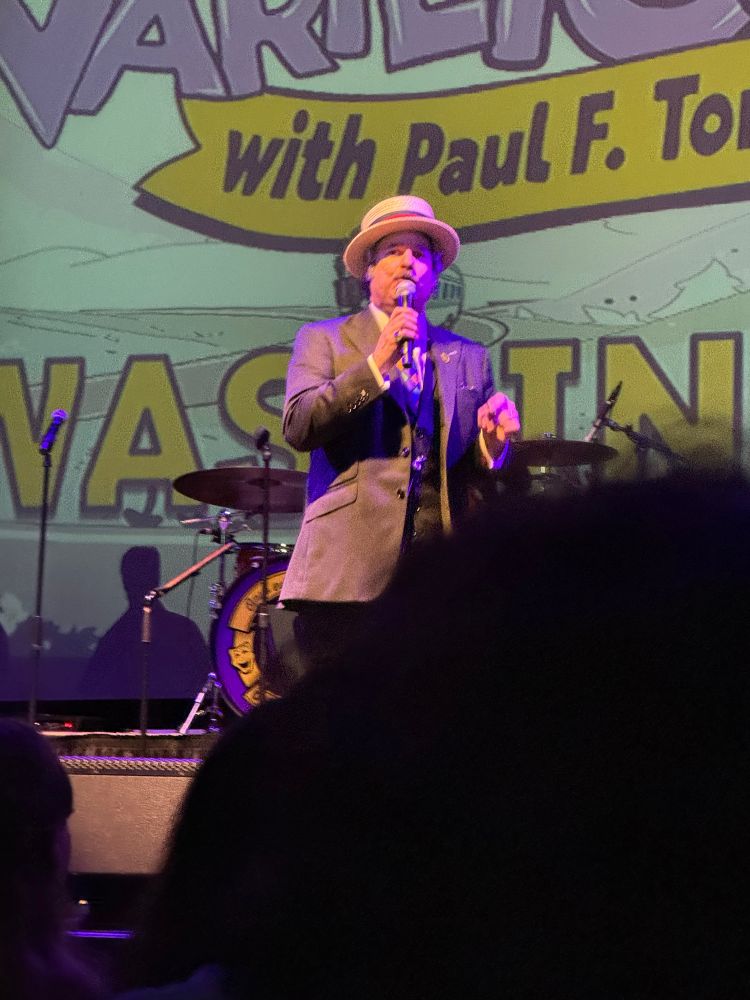 Paul F Tompkins on stage at Varietopia, wearing a suit and old-timey straw hat standing in front of an elevated drum kit, with a large display screen in the background with the Varietopia logo