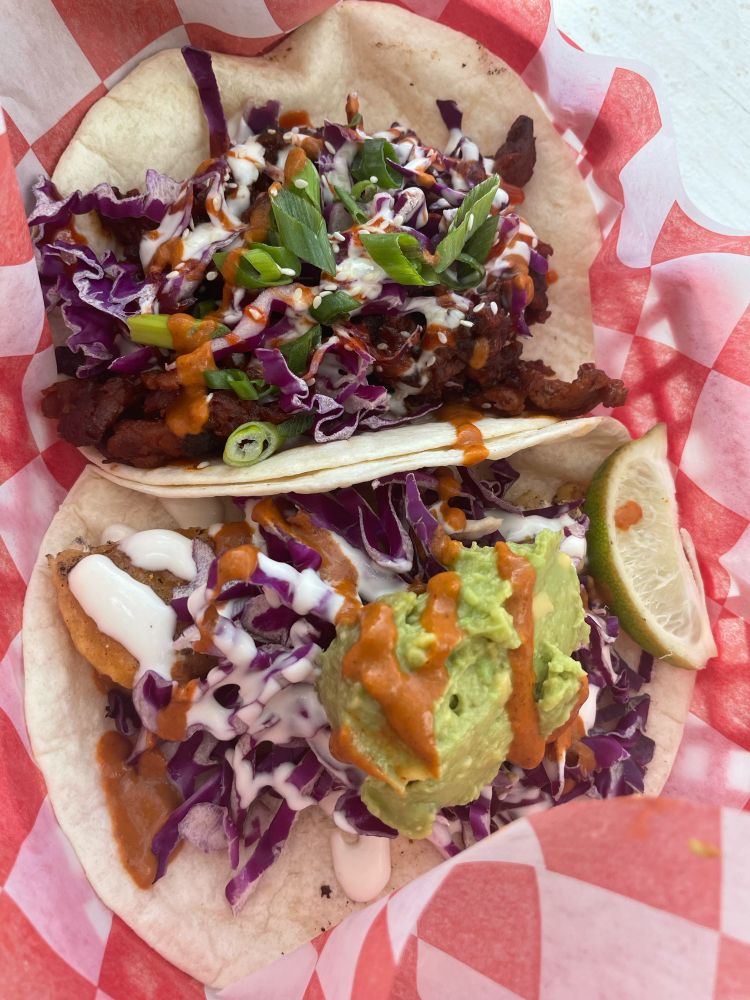 Two fat vegan tacos in a to-go basket with red and white checkered paper from the Vegan Nom food truck. One taco is Korean BBQ, and the other is a Del Rey fishless taco with a lime wedge garnish