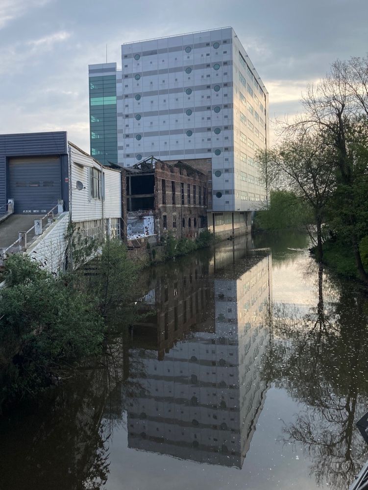 Portrait format landscape photo showing a modern riverside building, a derelict brick workshop and a modern warehouse. They're all reflected in a slow-moving river in morning light. 