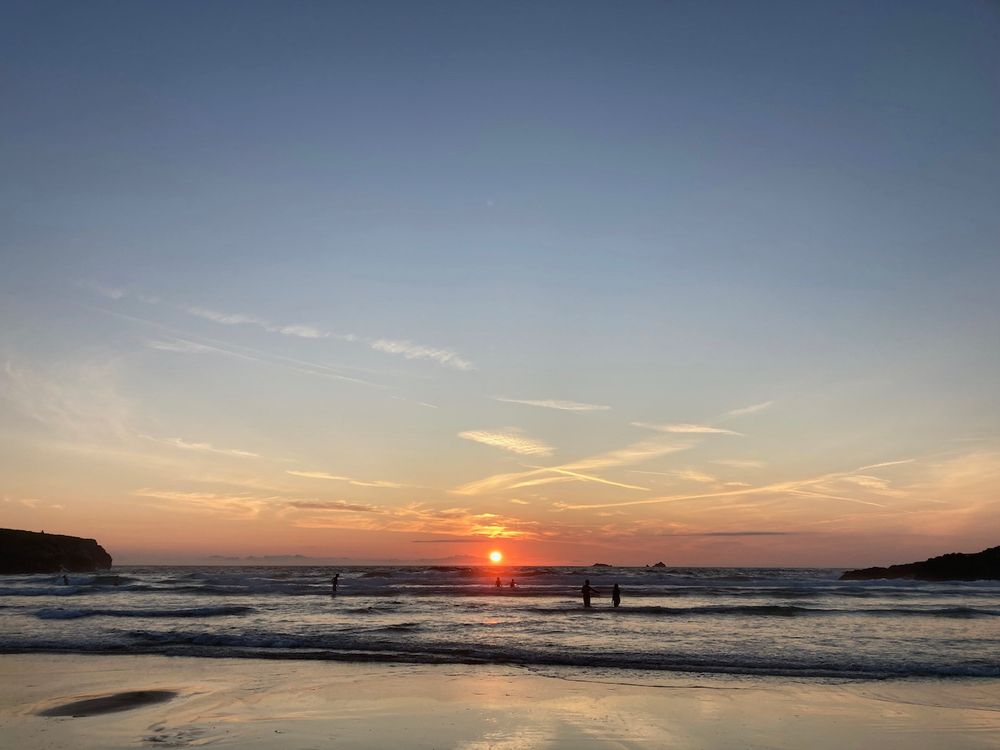 Landscape format photo of a beach at sunset with a few figures in the waves silhouetted against the light