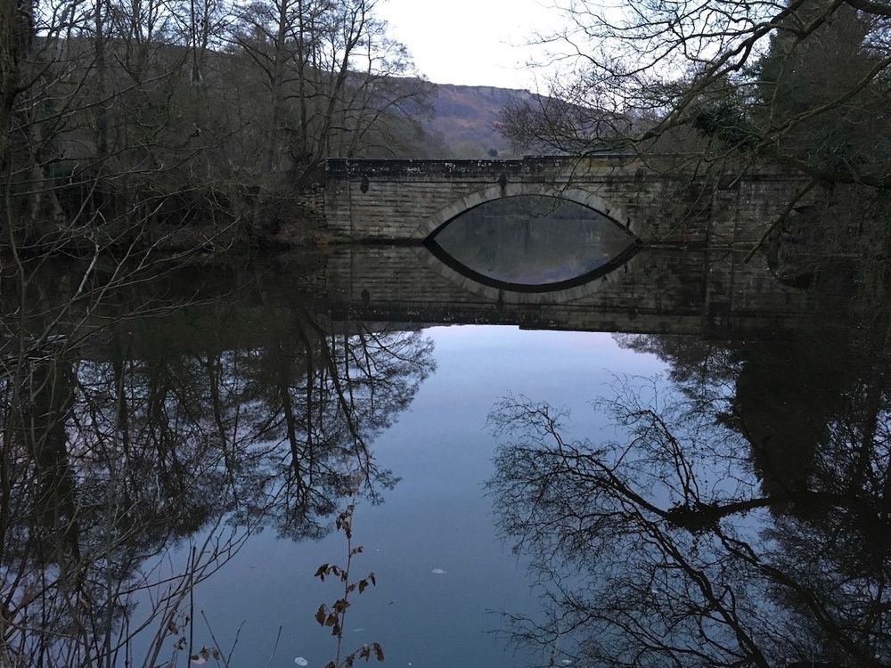 Landscape photo of a stone bridge and surrounding trees reflected in a slow moving river. A gritstone edge is visible in the background. The bridge and its reflection together make the shape of an eye. 