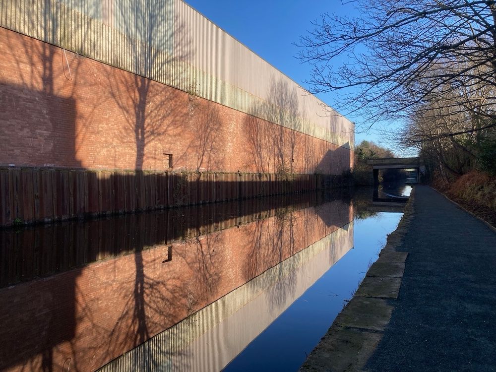 Landscape format photo of a line of winter tree shadows cast onto a long brick warehouse and reflected in the still water of a  canal which leads into the distance. 