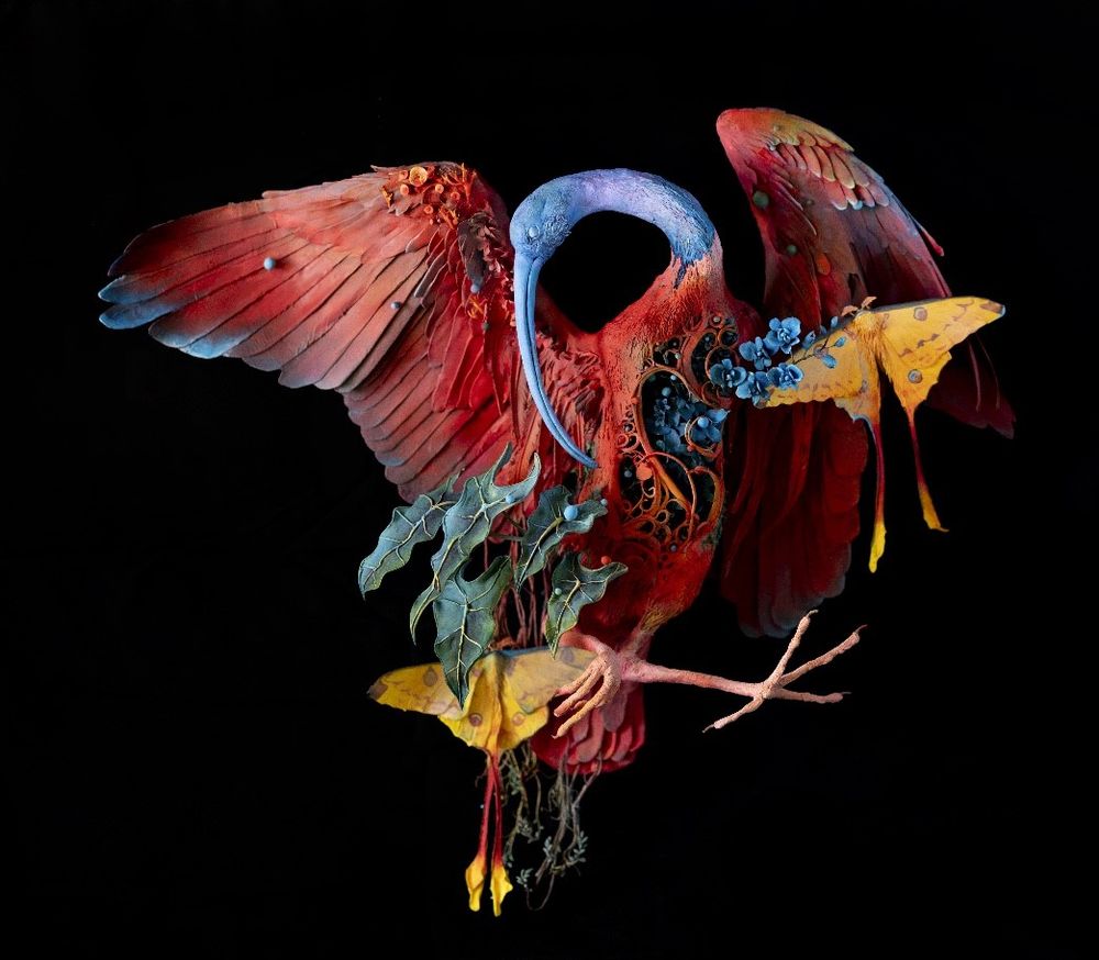 A surrealist sculpture featuring a blue, red, and orange ibis in flight with its head turned to the side. There are blue flowers sprouting from its chest with yellow luna moths and alocasia leaves.