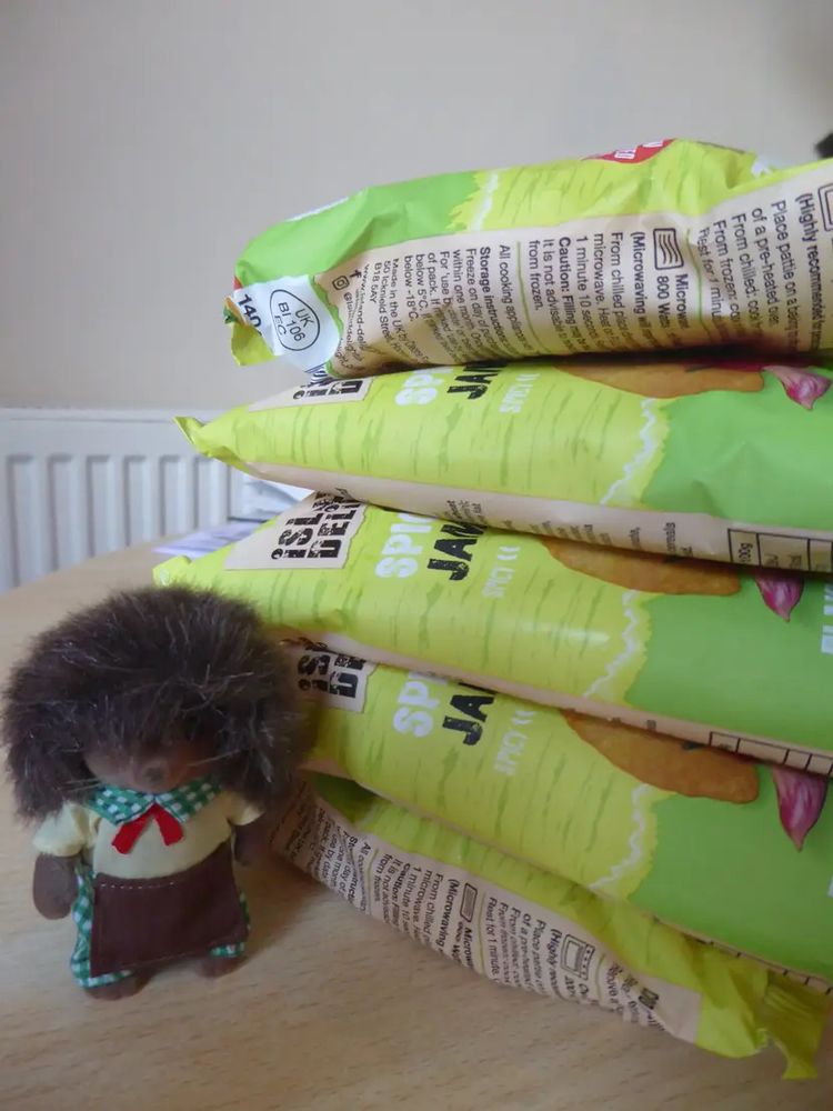 Five vegetable Jamaican patties in their packets in a pile on a plain table with a Sylvanian hedgehog pizza dad next to them. He is extremely furry and is wearing a yellow top with red necktie over a green & white checked shirt & trousers, plus a brown apron - kinda “Italian rustic” I guess.