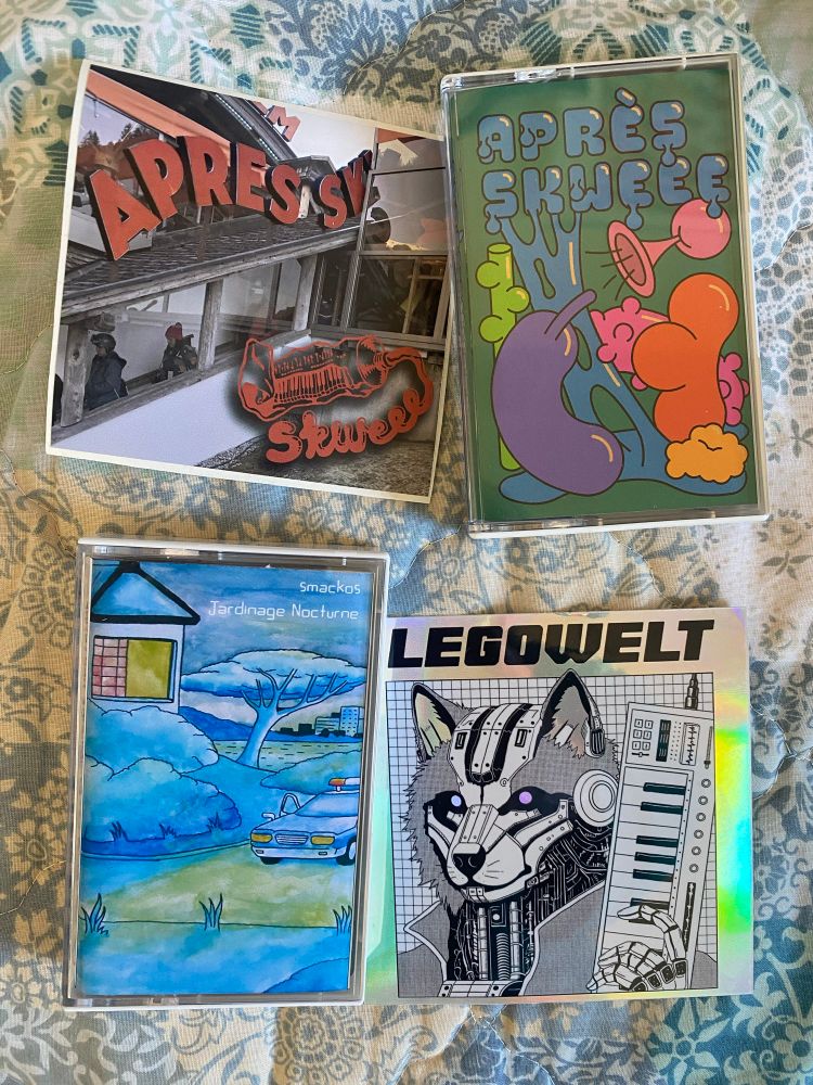 Two cassette tapes with nice colourful artwork, against a floral fabric, each tape also has a sticker next to it. One tape is called Apres Skweee & has a sticker with the “famous” skweee toothpaste tube logo, the other tape is Jardinage Nocturne by Smackos, the sticker is reflective (hologram) silver with text saying Legowelt, with a cool cyborg wolf holding a small synth (or keyboard controller) like a gun.