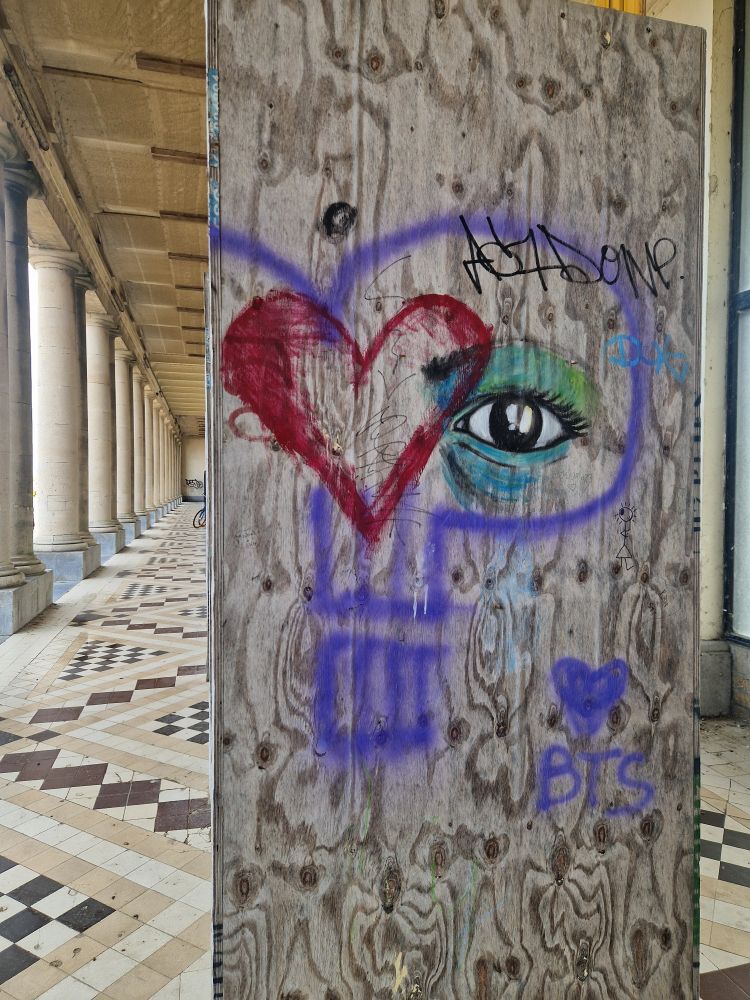 Tiled seaside arcade under repair - the wooden casing of a column has been graffitied, and there is a '💜BTS' - armi is everywhere...