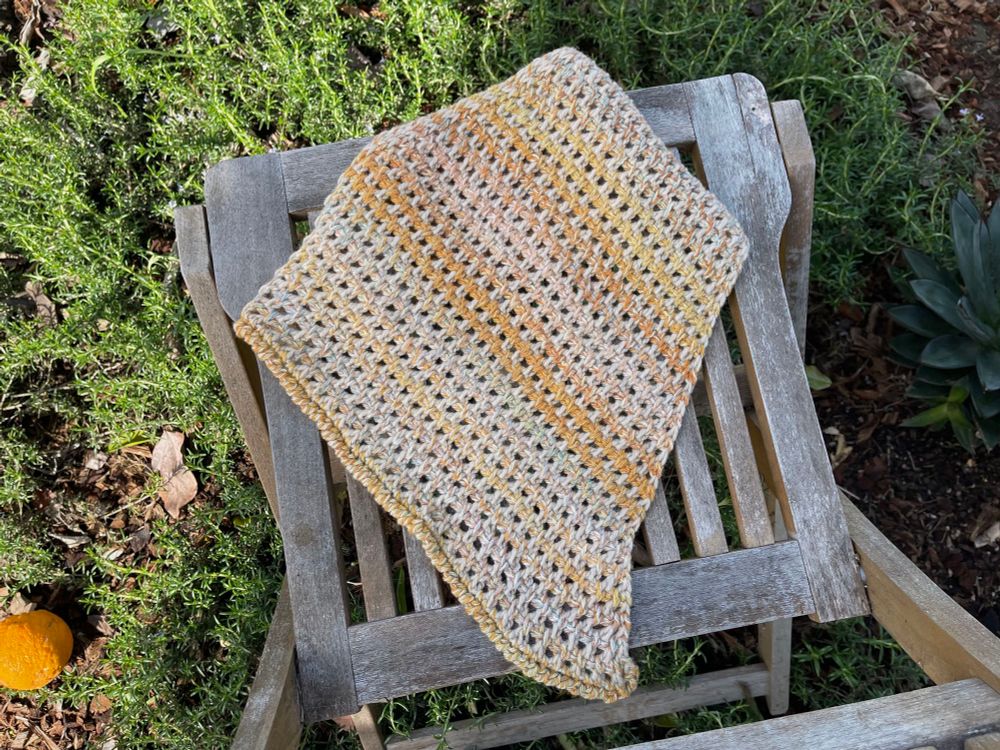 A cowl crocheted out of a mixture of beige yarn and variegated handspun yarn is placed on top of an outdoor chair. The handspun yarn changes colors in between different shades of orange and light blue. The chair is sitting on a garden of rosemaries, with an orange dropped to the ground and blue agave peeking in the sides of the picture.  