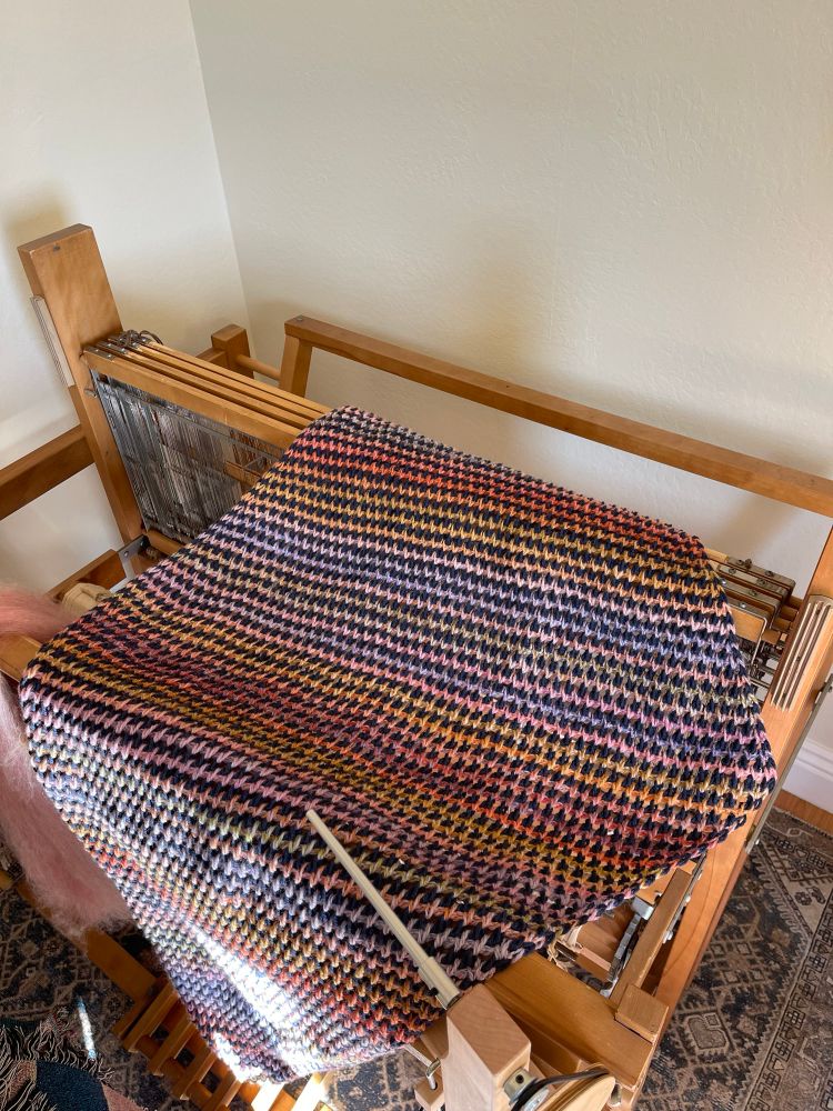 A big triangular shawl spread over a floor loom. The shawl is comprised of solid navy and variegated colors.  