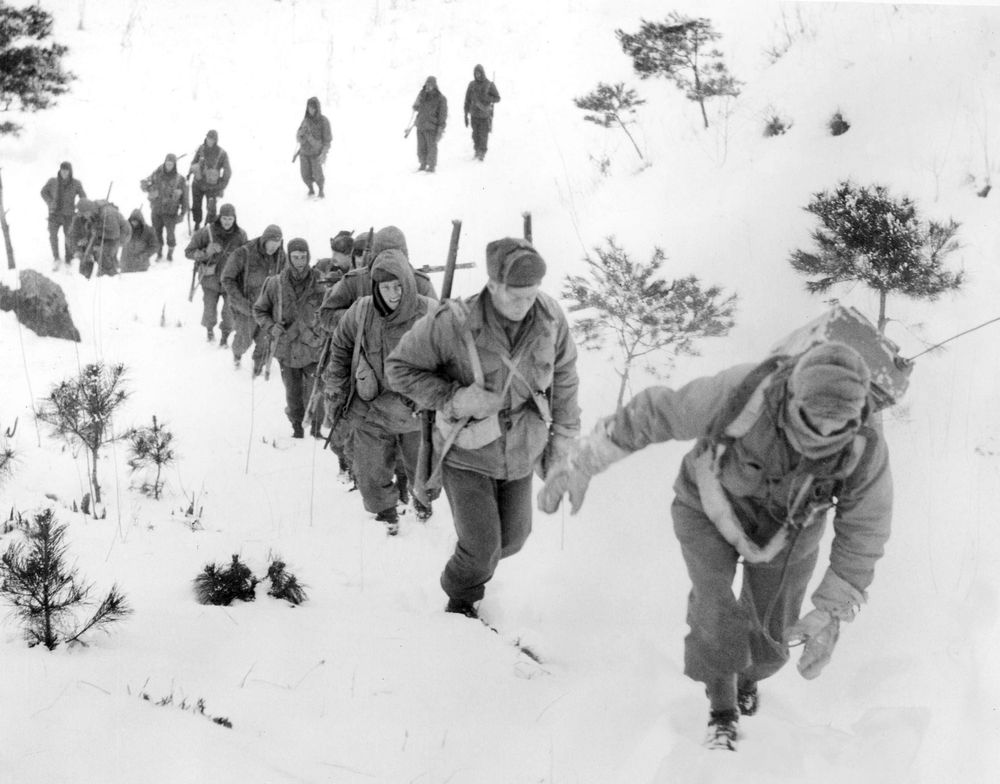 Soldiers on patrol in the Korean War in 1951 trudge through snow, through which the tops of bushes emerge. The photo is in black and white.